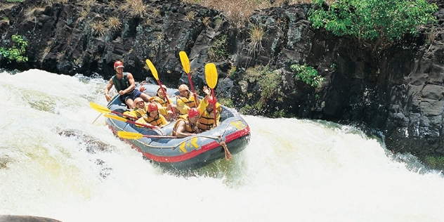 a group of people wearing life jackets and hard hats huddle in an inflatable dinghy steered by their guide as it speeds down river rapids