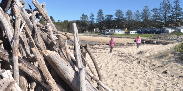 large pile of driftwood stacked for a beach bonfire with two children running along the beach in the background