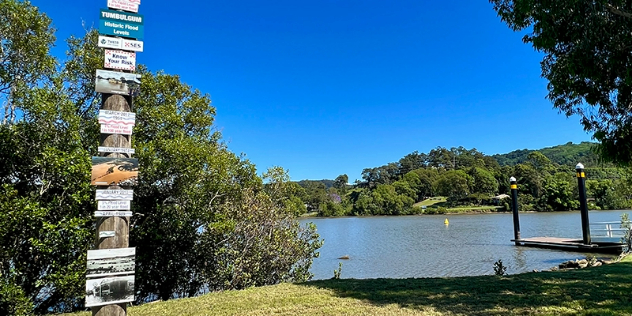  A sunny, treed riverbank, with a pier and a wood post on shore with historical information, photos and swim warnings.