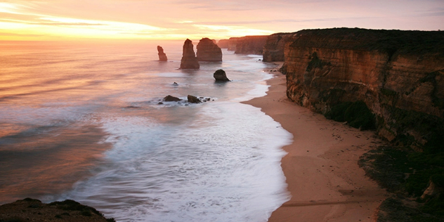 Rocky cliffs lead to a sand beach and sea with four of the twelve apostle rock formations visible, bathed in warm orange sunset.