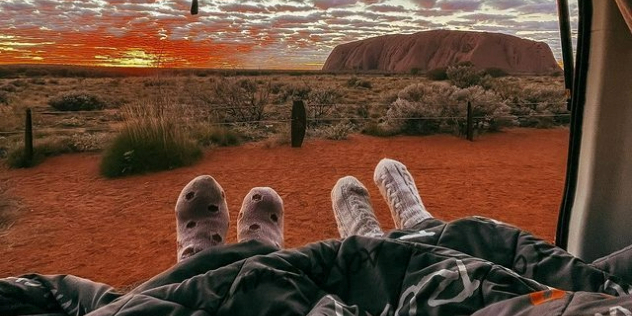 A sunset at the Uluru rock formation from the perspective of two people laying in a hatchback covered in a blanket.