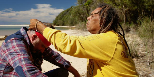  An Aboriginal Elder with dreadlocks, Uncle Micklo, puts a bandanna over the head of a bearded man on a sunny beach.