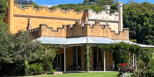 A two level colonial style house, painted in a warm orange with brown trim, with a four poster veranda opening onto grass, hugged by trees under a bright blue sky.