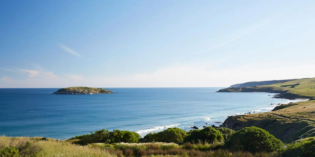 Green fields flow down to a rocky shoreline and large deep blue bay, with a single small green island under blue sky.