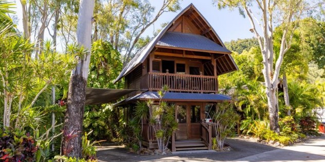 A two story, dark wood villa, surrounded by trees, with a veranda on the second level and porch below. A flat hammock is tied off the side of the veranda onto a nearby tree.