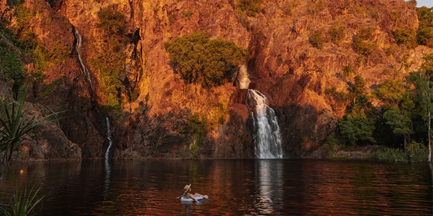 A waterfall falling into a pool of water from a craggy, scrubby hillside lit by orange sunset rays, a woman in a hat floats in a tube in the pool near foreground.