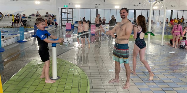 A boy in shorts and a swim shirt squirts a man with a goatee in shorts with a water gun, in a busy, indoor pool and waterpark.