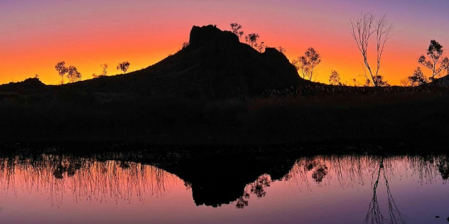 A pond perfectly mirrors the silhouette of a hillside against a purple, orange and yellow sunset.