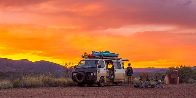 a woman stands next to her camper van at a lookout admiring the vivid orange and yellow sunset over distant hills