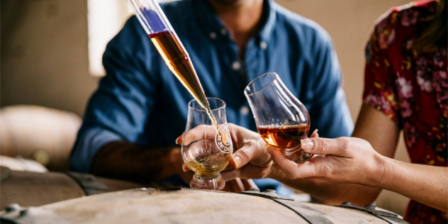 A man and woman lean over wooden barrels, holding out glasses. The man’s is being filled by a long glass pipette filled with carmel-coloured whiskey. 