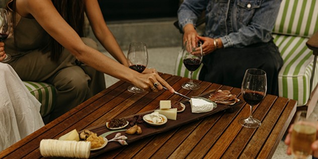 Two women and a man sit around a wood-plank table with glasses of red wine, eating from a dark wood serving board, covered with cheeses. 