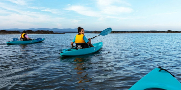 A woman canoeing near a treed shore and small islands smiles over her shoulder at the camera.