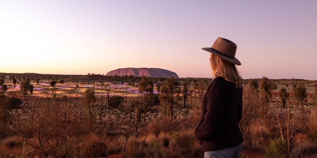  A woman in an Australian wide brim hat looks out over glowing yellow and purple lights at the base of the Uluru rock formation.