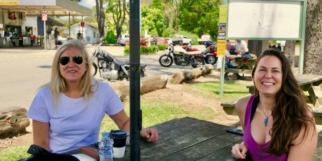  A blond and a brunette woman smile while sitting at a picnic table outside a cafe and petrol station with motorcycles out front.