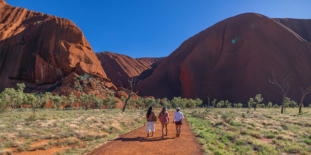 Three women in sunhats seen from behind as they walk up to the base of the Uluru rock formation along an cleared path of orange dirt, surrounded by green scrub under a clear blue sky. 