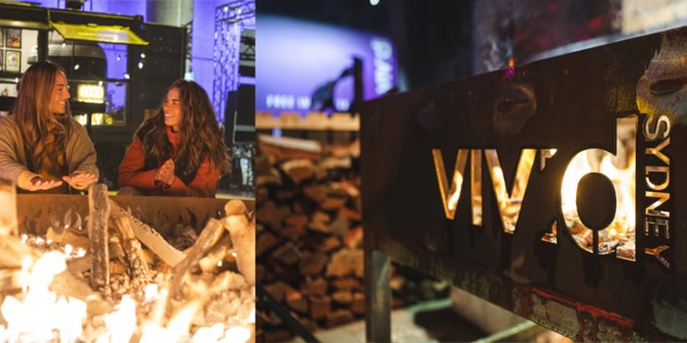  Two women smile and warm themselves by an outdoor fire, next to a metal Vivid sign, backlit by the fire.