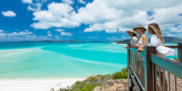 Three women on a balcony along a rocky cliff look over a vista of white sandbars and shallow turquoise water, leading to treed islands in the distance. 