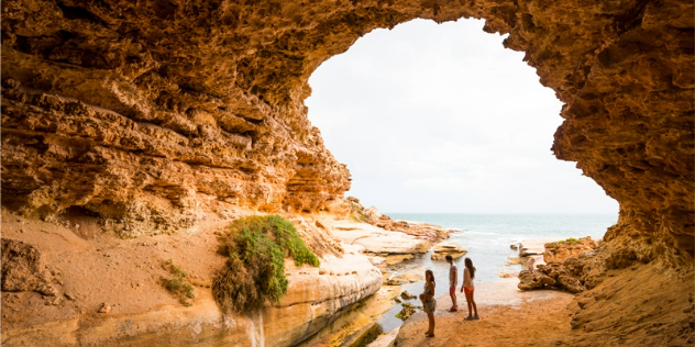 3.	The mouth of a large ocean rock cave framing the sea, as seen from inside on an overcast day. Its entrance towers about 15 metres over three teens standing at the base of the cave.