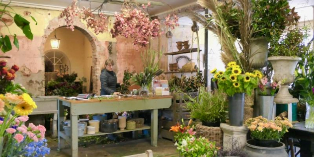 A bench with an stone arch behind it, inside a florist and cafe, all surrounded by flowers in bouquets and pots. 