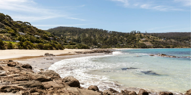  Rocks leading to a rounded, sandy beach with green hills rising around it dotted with houses.