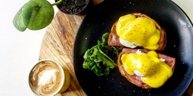 close up of eggs benedict and garnish on a black plate and a cappuccino 